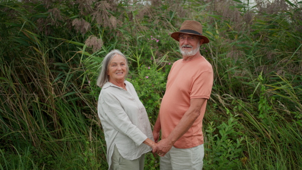 Happy senior couple holding hands, smiling at each other and looking into camera. Green river plants in background.