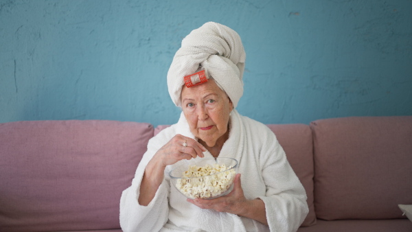 Happy senior woman sitting at a sofa in bathrobes and watching TV with popcorn.