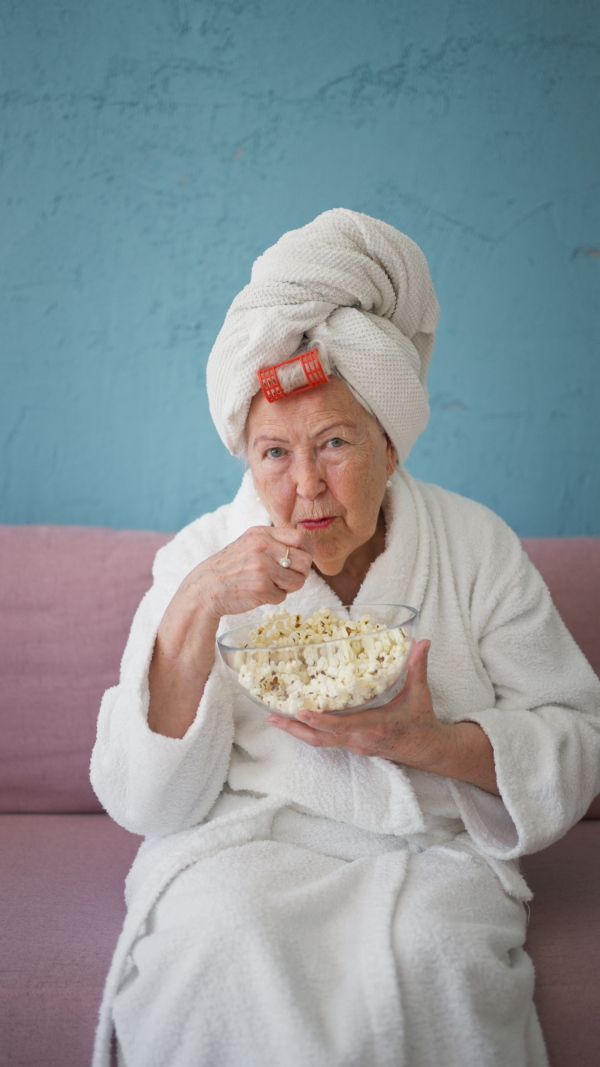 Happy senior woman sitting at a sofa in bathrobes and watching TV with popcorn.