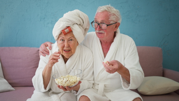 Happy senior couple sitting at a sofa in bathrobes and watching TV with popcorn.