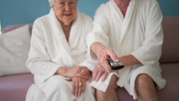 Happy senior couple sitting at a sofa in bathrobes and watching TV.