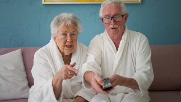 Happy senior couple sitting at a sofa in bathrobes and watching TV.