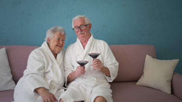 Happy senior couple sitting together in the bathrobe on sofa with glass of wine, having nice time at home.