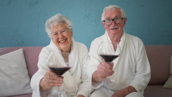 Happy senior couple sitting together in the bathrobe on sofa with glass of wine, having nice time at home.