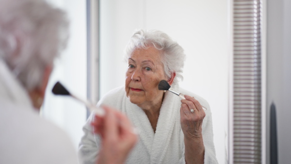 Senior woman standing in front of mirror in her bathroom and preparing her face with make up brush.