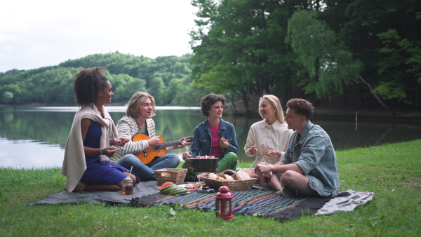 A group of young friends having fun on picnic near a lake, sitting on blanket eating and playing guitar.