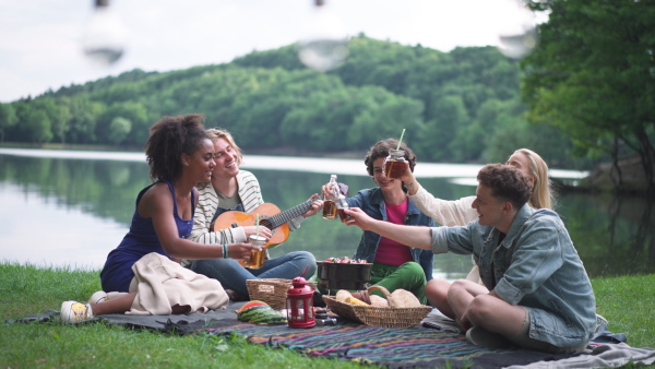 A group of young friends having fun on picnic near a lake, sitting on blanket eating and playing guitar.