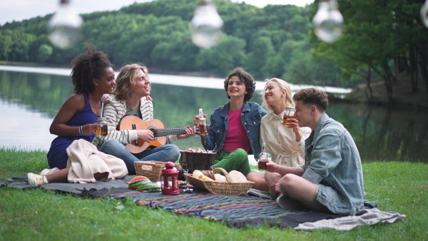 A group of young friends having fun on picnic near a lake, sitting on blanket eating and playing guitar.