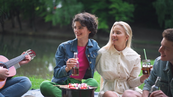 A group of young friends having fun on picnic near a lake, sitting on blanket eating and playing guitar.