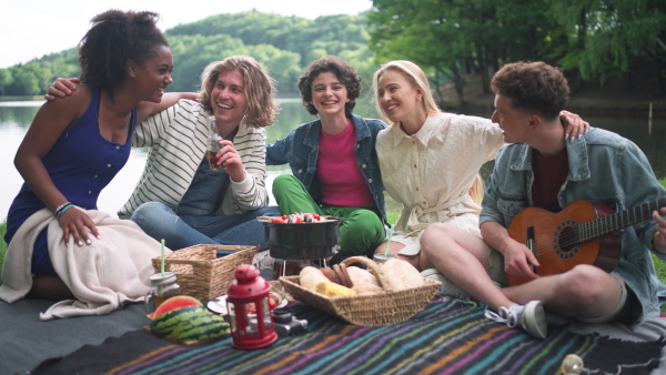 A group of young friends having fun on picnic near a lake, sitting on blanket eating and playing guitar.