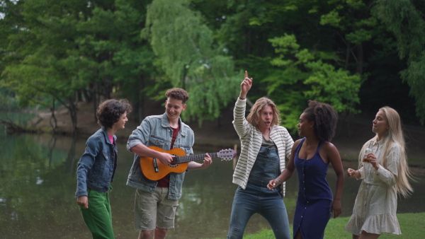 A group of young friends having fun on picnic near a lake, dancing, singing and playing guitar.