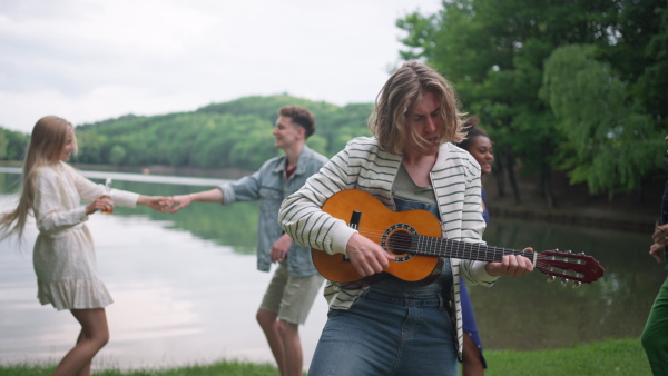 A group of young friends having fun on picnic near a lake, dancing, singing and playing guitar.