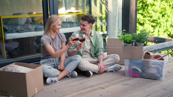 A cheerful young couple moving in their new tiny house in woods sitting on floor on terrace and drinking wine. Conception of moving and sustainable living.