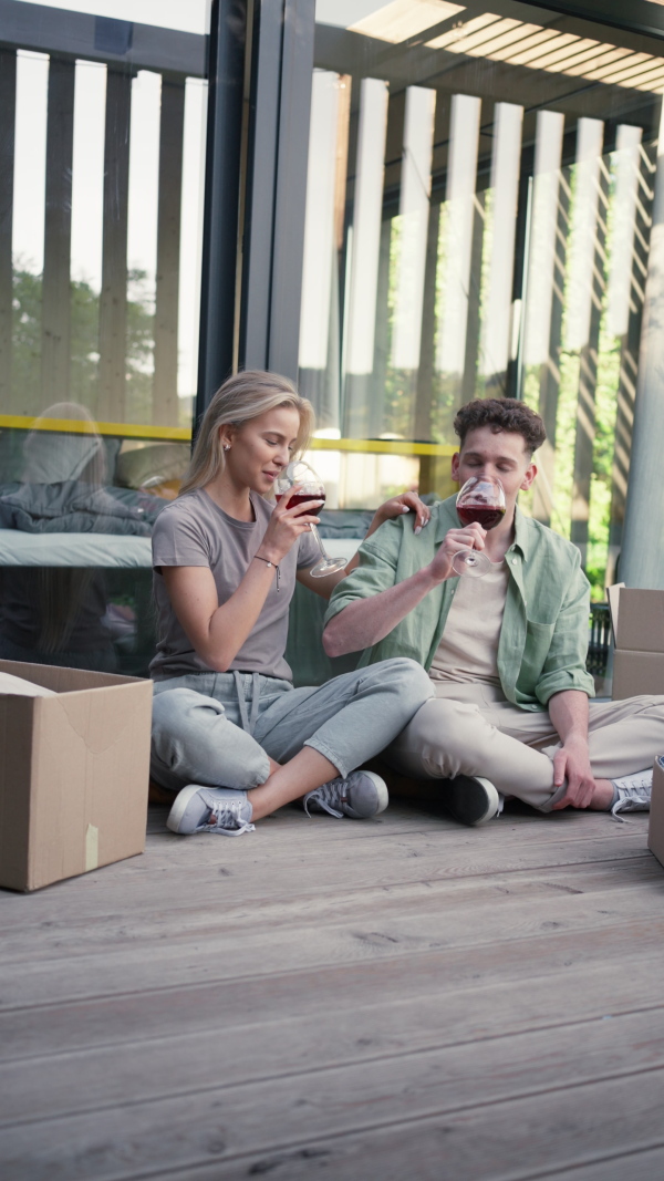 A cheerful young couple moving in their new tiny house in woods sitting on floor on terrace and drinking wine. Conception of moving and sustainable living.