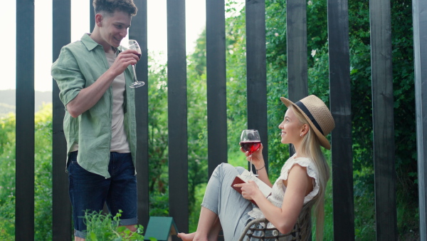 A young beautiful woman relaxing on cozy terrace, reading a book, her boyfriend is bringing her glass of wine, digital detox and weekend away concept.