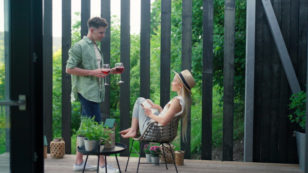 A young beautiful woman relaxing on cozy terrace, reading a book, her boyfriend is bringing her glass of wine, digital detox and weekend away concept.