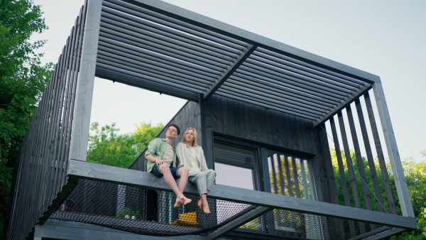 A young couple sitting and cuddling in hammock terrace in their new home in tiny house in woods, sustainable living concept.