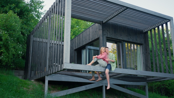 A young couple sitting and cuddling in hammock terrace in their new home in tiny house in woods, sustainable living concept.