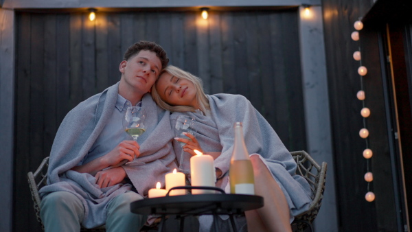 A young couple with wine resting outdoors on terrace in evening, weekend away in tiny house in countryside, sustainable living.