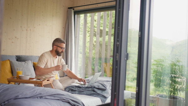 A mature man having breakfast and working in tiny house, in front of nature. Freelancer office concept