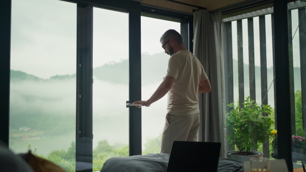 A mature man standing in tiny house terrace in front of beautiful nature with cup of coffee. Morning routine.