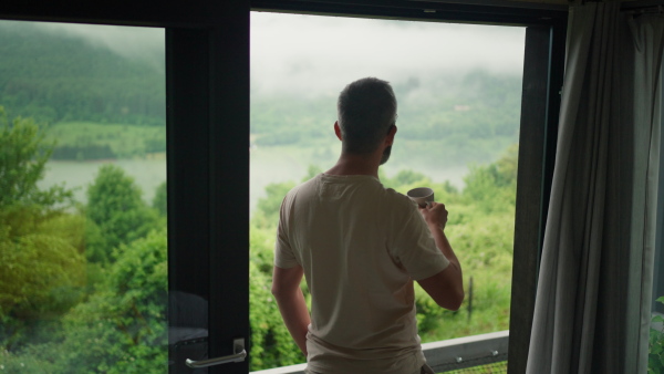 Back view of man in the morning light, standing at window, drinking coffee, looking at wonderful nature.