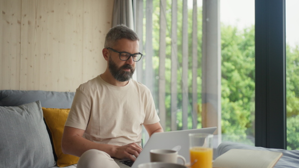 Maure man working on laptop in bed, while having breakfast, in room with nature view.