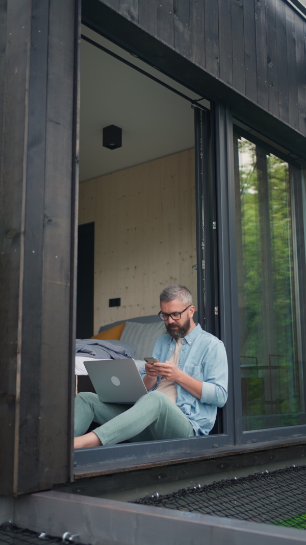 A mature man using laptop and working in tiny house, in front of nature. Freelancer office concept