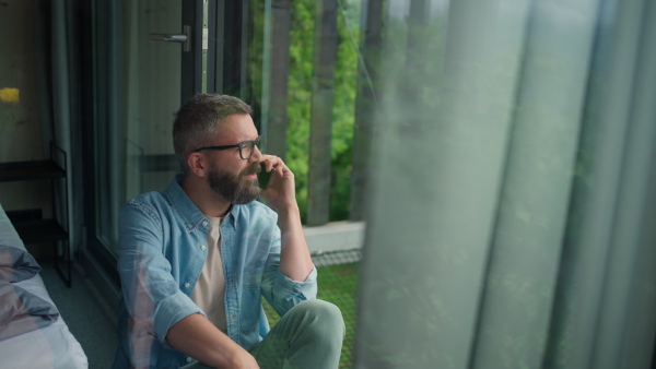 Mature Man sitting on floor by a window at home, talking on phone, enjoying the view in nature