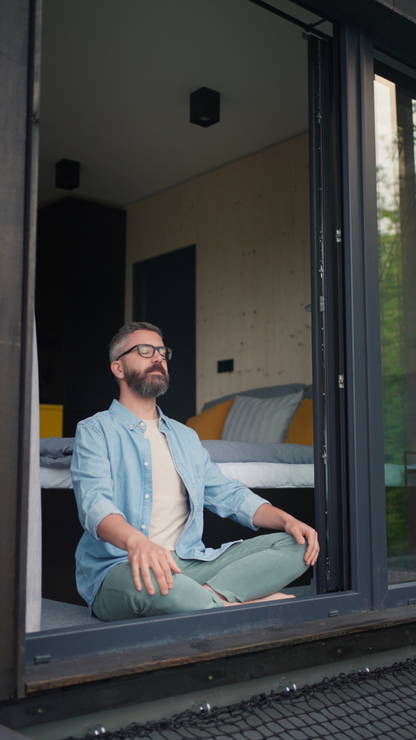 Mature man meditating at open terrace window, breathing deeply. Morning routine.