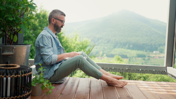 Freelancer working on his laptop, on a terrace of a cabin in the woods, surrounded by beautiful nature