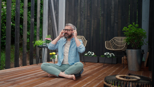 A mature man sitting in terrace, listening music with headphones and enjoying summer time.