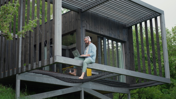 Mature man sitting on modern cabin terrace working on laptop, listening to music