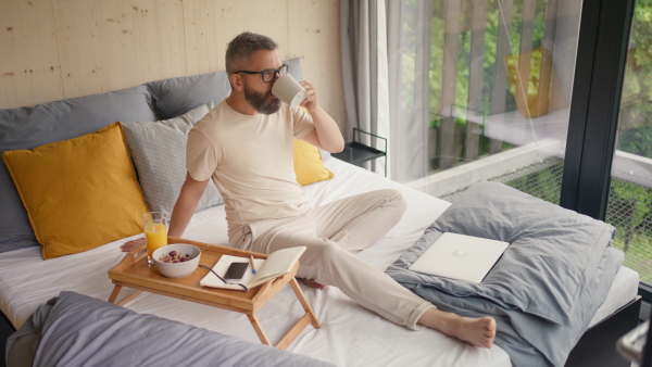 Mature man having breakfast and drinking coffee in sustainable tiny house, in front of nature