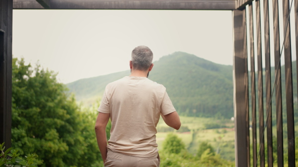 Back view of mature man standing on cabin terrace, drinking coffee, enjoying beautiful nature view.