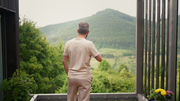 A mature man standing in tiny house terrace in front of beautiful nature with cup of coffee. Morning routine.