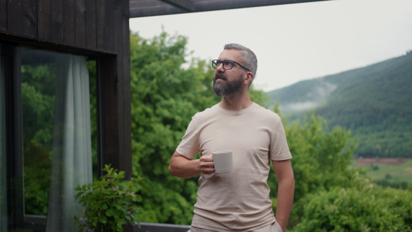 A mature man standing in tiny house terrace in front of beautiful nature with cup of coffee. Morning routine.