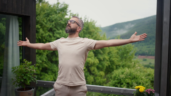 Mature man standing on terrace in nature, outstreched arms, breathing deeply, connecting to nature, enjoying.