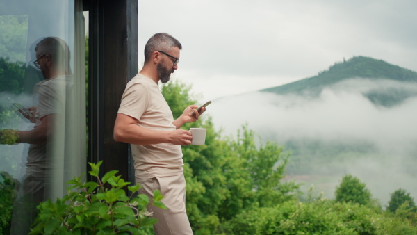 Mature man standing on terrace in beautiful nature in the morning, looking at his phone with cup of coffee.