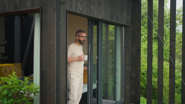 A mature man standing in tiny house terrace in front of beautiful nature with cup of coffee. Morning routine.