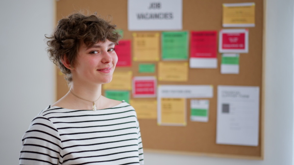 A happy young woman student standing in front of employment noticeboard, looking at camera.