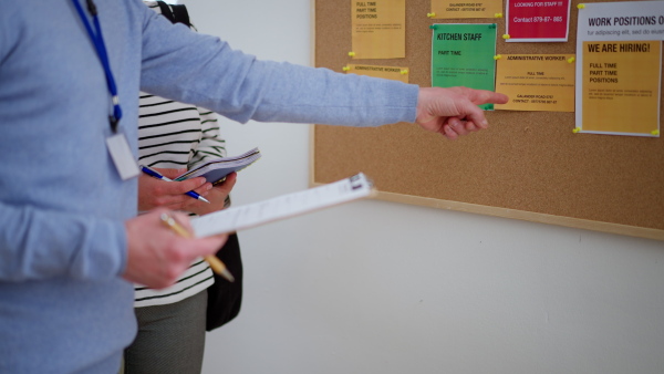 A recrutiment agency employee standing in front of employment noticeboard and helping young woman to search for a job, close-up.