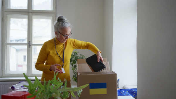 A senior volunteer woman packing box with Humanitarian aid for Ukrainian refugees in office