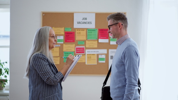 A recrutiment agency employee standing in front of employment noticeboard and helping mature man search for a job.