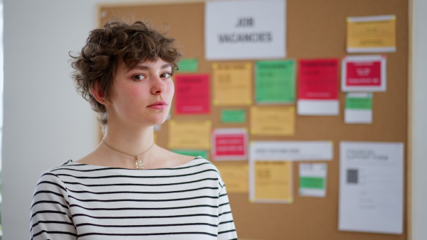 A happy young woman student standing in front of employment noticeboard, looking at camera.