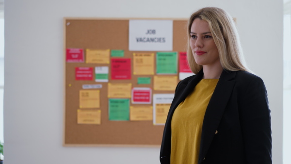 A happy young woman student standing in front of employment noticeboard, looking at camera.