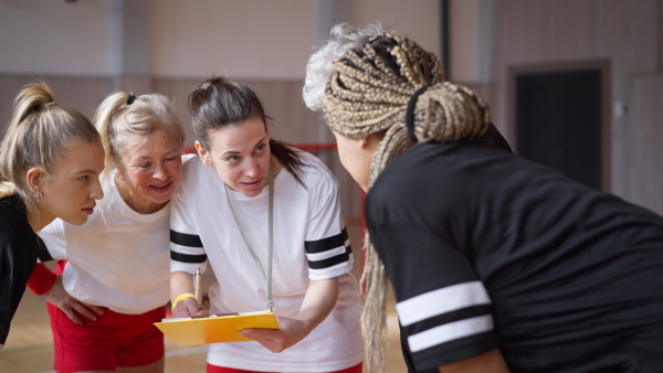 A female sport coach with clipboard discussing tactics with young and old women team training for match in gym.