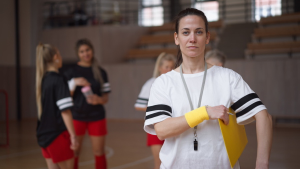 A female sport coach with clipboard looking at camera with young and old women team on training in gym at background.