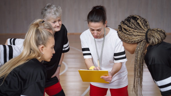 A female sport coach with clipboard discussing tactics with young and old women team training for match in gym.