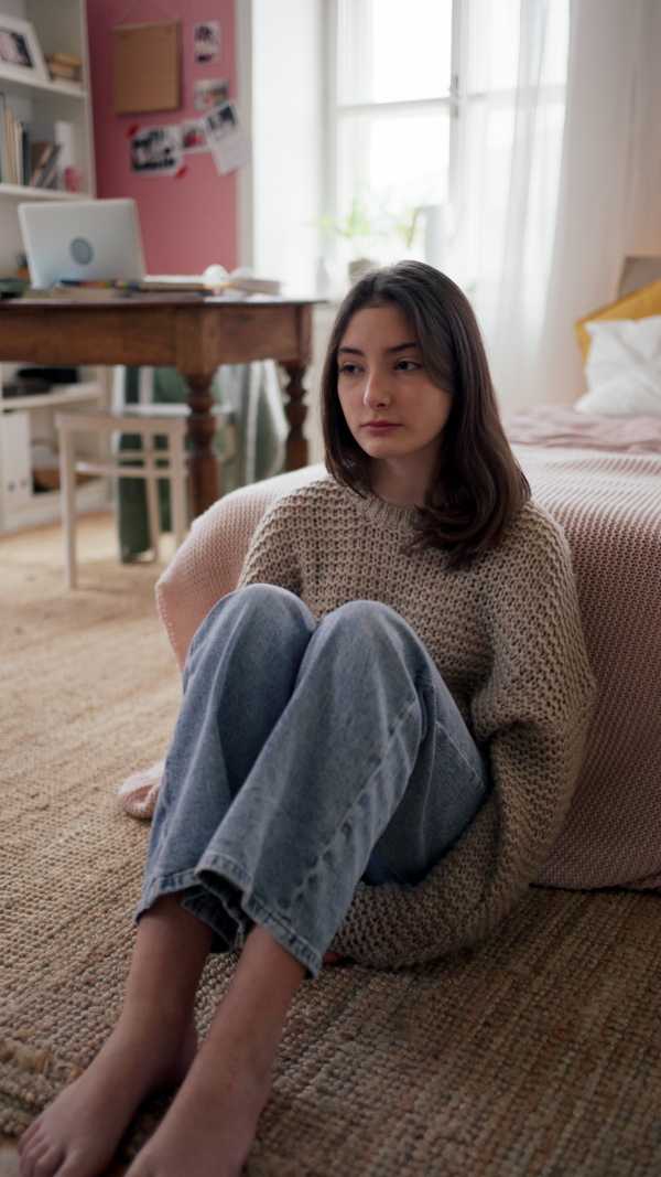 Vertical view of a sad teenage girl sitting on the floor in her room.